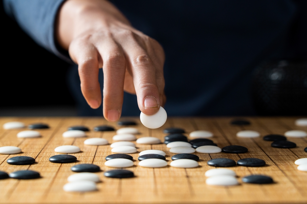 Illustration des relations entre management et syndicats à travers le jeu de Go : vue rapprochée d'une main posant une pierre blanche sur un plateau en bois où coexistent de nombreuses pierres noires et blanches.