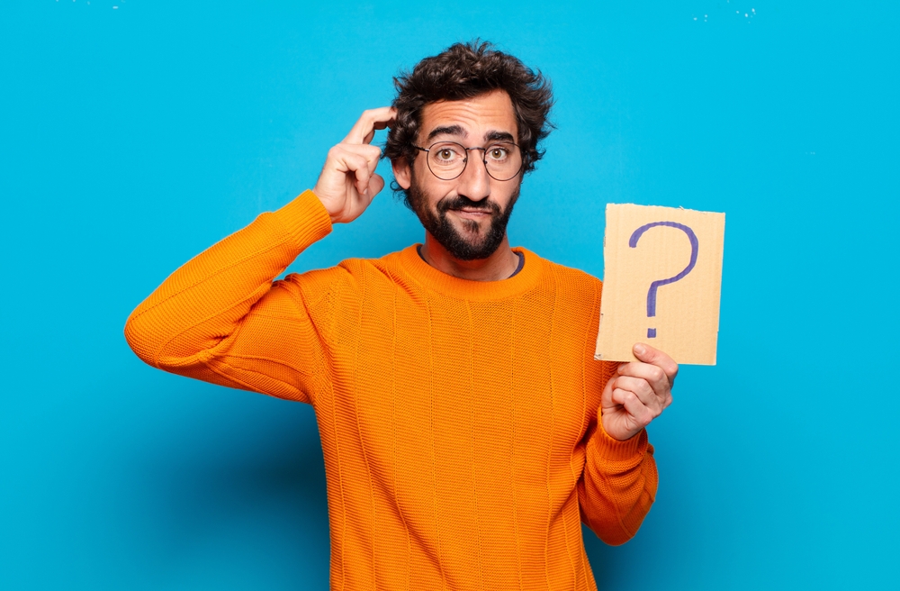 Un homme au pull orange et aux lunettes gratte sa tête avec un air perplexe tout en tenant un panneau en carton avec un point d'interrogation, symbolisant la confusion ou une interrogation.