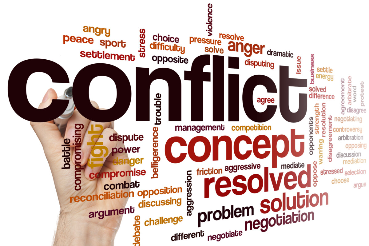 Nuage de mots en anglais autour du thème du conflit, avec des termes tels que 'conflict', 'resolved', 'concept', 'solution', 'negotiation', 'fight', 'argument', 'mediation' et d'autres mots-clés relatifs aux tensions, à la résolution, à la violence et à la réconciliation. Une main écrit le mot 'conflict' au marqueur noir sur un fond blanc.