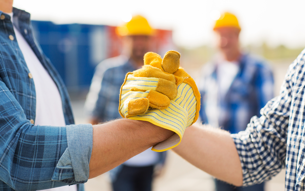 Deux travailleurs en tenue de chantier, portant des chemises à carreaux, se serrent la main fermement, symbolisant la coopération et la solidarité. L’un porte des gants de protection jaunes. En arrière-plan flou, deux autres ouvriers avec des casques de sécurité jaunes observent la scène sur un chantier. L'image évoque la confiance, l'entraide et la collaboration dans le milieu professionnel.