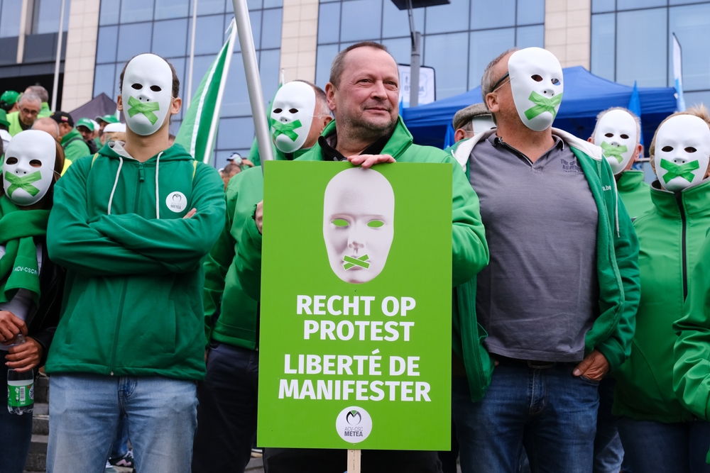 Des manifestants vêtus de vert, certains portant des masques blancs barrés d’un ruban vert sur la bouche, participent à une action de protestation. Au centre, un homme sans masque tient une pancarte verte sur laquelle est écrit en néerlandais et en français : « Recht op protest / Liberté de manifester ». Le groupe est rassemblé devant un bâtiment moderne, exprimant ainsi une revendication pour le droit à la liberté d’expression et de manifestation.