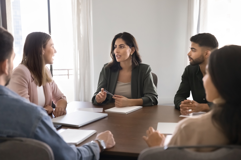 Un groupe de cinq personnes en réunion autour d’une table dans une salle lumineuse. Une femme au centre, tenant un stylo, semble mener la discussion tandis que les autres participants, hommes et femmes, l’écoutent attentivement. Des carnets et des ordinateurs portables sont posés devant eux. L’image illustre un travail d’équipe, une séance de brainstorming ou une prise de décision collaborative en entreprise.