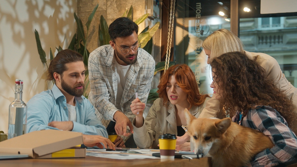 Cinq jeunes adultes, hommes et femmes, collaborent activement autour d’une table dans un espace de travail décontracté et lumineux. Ils examinent des documents avec concentration et discussion. Un chien de type corgi est assis calmement sur les genoux d’une participante, ajoutant une touche conviviale à l’ambiance. Des objets du quotidien, comme une bouteille, une boîte en carton et un café à emporter, sont visibles sur la table. L’atmosphère est dynamique et créative, typique d’un environnement de coworking ou d’une startup.