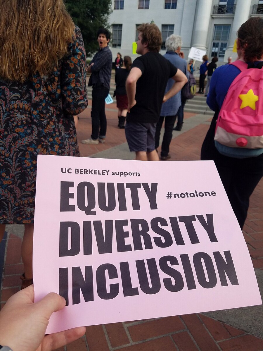 Une personne tient une pancarte rose lors d’un rassemblement devant un bâtiment universitaire, sur laquelle on peut lire « UC Berkeley supports Equity, Diversity, Inclusion #notalone ». Cette image illustre la mobilisation pour l’équité, la diversité et l’inclusion, des valeurs essentielles pour prévenir et résoudre tout conflit en entreprise, notamment en lien avec les discriminations, l’exclusion ou les inégalités de traitement.
