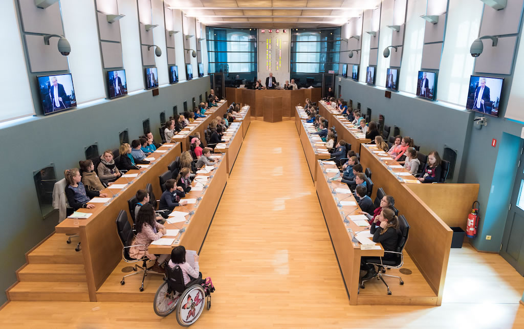 Vue intérieure de la salle du parlement wallon rempli d’enfants ou de jeunes assis à des pupitres organisés en deux rangées parallèles. Au fond de la pièce, deux adultes se tiennent derrière un bureau surélevé, évoquant un rôle de présidence ou de modération. Plusieurs écrans muraux diffusent l’image d’un homme en costume, suggérant une session retransmise ou enregistrée. L’environnement est lumineux, moderne et inclusif, avec la présence visible d'une jeune fille en fauteuil roulant. L’ambiance évoque un projet pédagogique de simulation parlementaire ou d’éducation citoyenne.