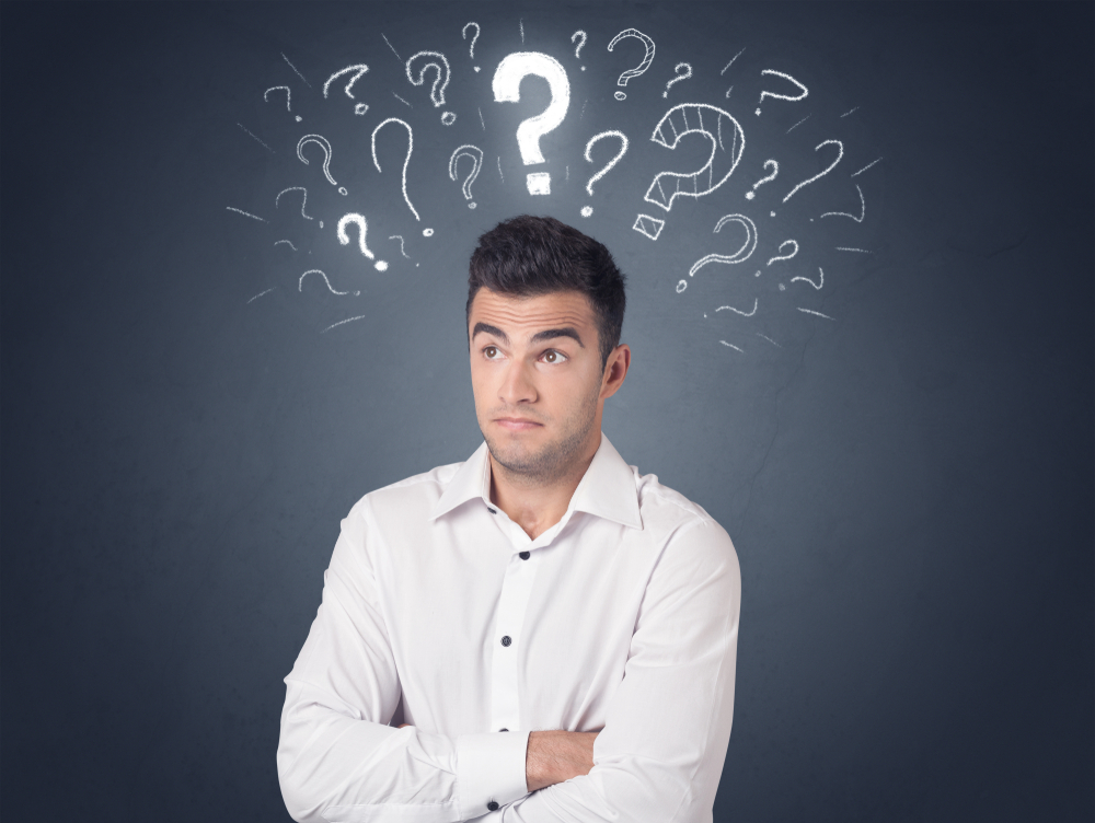 Un homme en chemise blanche, les bras croisés, regarde vers le haut avec une expression interrogative sur le visage. Au-dessus de sa tête, plusieurs points d’interrogation dessinés à la craie flottent sur un fond ardoise, symbolisant la confusion, le doute ou une intense réflexion. L’image évoque l’incertitude ou le questionnement face à une décision à prendre ou un problème à résoudre.