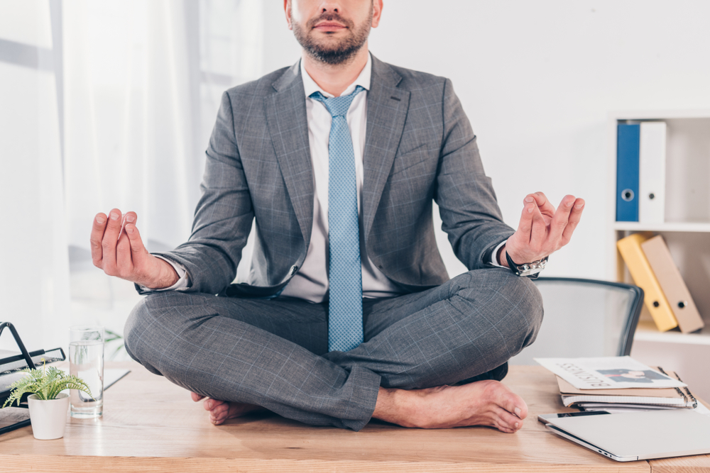 Un homme en costume-cravate est assis en tailleur sur un bureau, les yeux fermés, les mains posées sur les genoux en position de méditation. Cette scène illustre une tentative de gestion du stress et de recherche d’équilibre intérieur, probablement en réponse à un conflit au travail, soulignant l'importance de la régulation émotionnelle dans un environnement professionnel tendu.