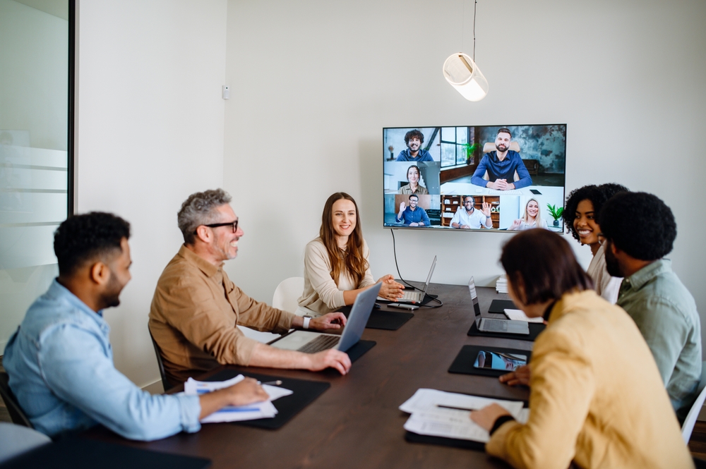 Réunion hybride dans une salle de conférence, où plusieurs collaborateurs sont réunis physiquement autour d’une table, tandis que d’autres participent à distance via visioconférence affichée sur un grand écran. Tous semblent engagés et souriants. Cette image illustre une dynamique de travail collaborative visant à prévenir ou à résoudre un conflit au travail grâce à une communication ouverte, inclusive et adaptée aux nouvelles formes d’organisation.