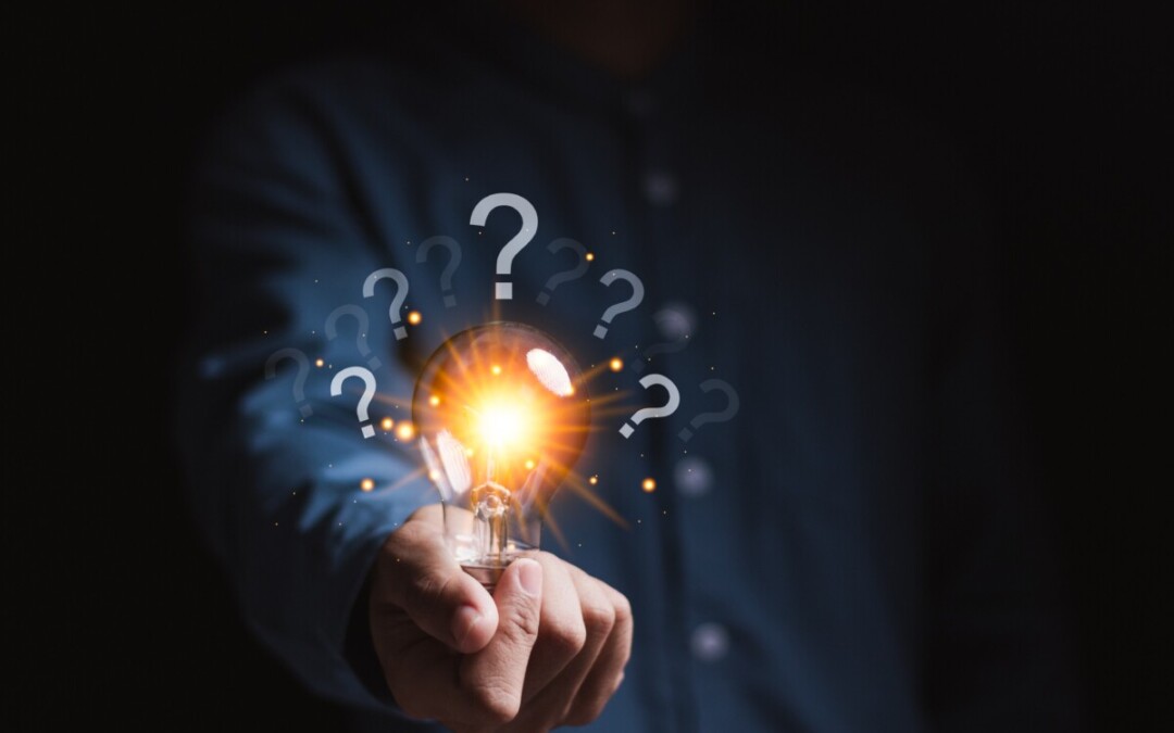 Une personne tend une ampoule allumée, tenue entre ses doigts, qui émet une lumière vive. Autour de l’ampoule, plusieurs points d’interrogation flottent, suggérant une réflexion, une recherche de solution ou une idée en émergence. L’arrière-plan sombre met en valeur la lumière et les symboles, renforçant l’idée d’un éclair de génie ou d’une réponse à une problématique complexe.