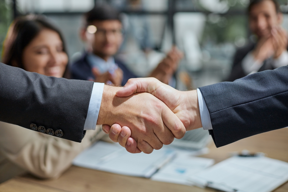Deux personnes en costume se serrent la main fermement au premier plan, symbolisant un accord ou la conclusion d’une négociation. En arrière-plan, des collègues souriants et applaudissant autour d’une table de réunion suggèrent un climat positif, professionnel et collaboratif. Des documents et un ordinateur portable sont posés sur la table, renforçant le contexte d’un accord formel ou d’une réussite collective en milieu de travail.