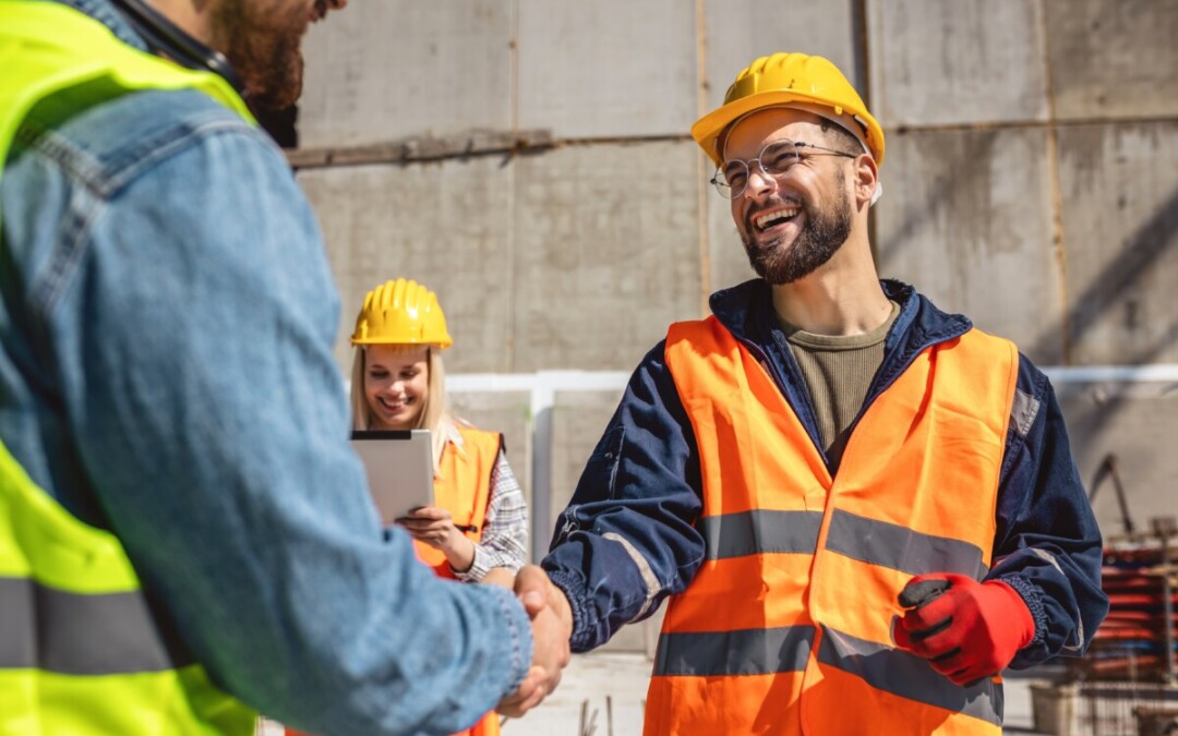 Sur un chantier, deux ouvriers portant des casques et des gilets de sécurité se serrent la main avec le sourire, tandis qu'une collègue en arrière-plan consulte une tablette. Cette image illustre une résolution positive d’un conflit au travail, mettant en avant l’importance du dialogue et de la coopération pour restaurer un climat de confiance au sein des équipes.