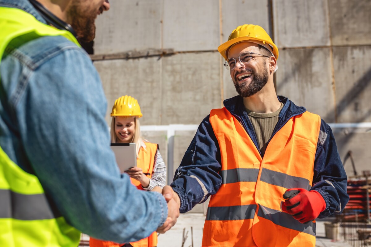 Sur un chantier, deux ouvriers portant des casques et des gilets de sécurité se serrent la main avec le sourire, tandis qu'une collègue en arrière-plan consulte une tablette. Cette image illustre une résolution positive d’un conflit au travail, mettant en avant l’importance du dialogue et de la coopération pour restaurer un climat de confiance au sein des équipes.