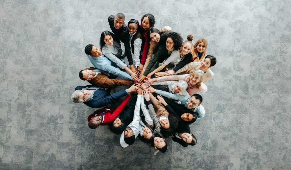 Photo prise en plongée d’un groupe diversifié de collègues formant un cercle et unissant leurs mains au centre dans un geste de solidarité. Cette image symbolise la résolution de conflit au travail par la coopération, l’inclusion et la cohésion d’équipe, illustrant l’importance de l’engagement collectif pour surmonter les tensions professionnelles.
