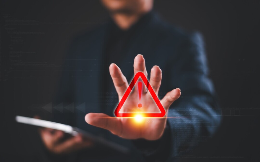 Un homme en costume, tenant une tablette, tend la main vers l’avant, où un symbole d’alerte rouge (triangle avec point d’exclamation) est superposé, évoquant un avertissement ou une situation critique. L’image est stylisée dans un environnement numérique sombre, suggérant un contexte de cybersécurité, de risque informatique ou de gestion de crise.