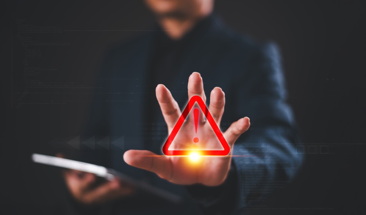 Un homme en costume, tenant une tablette, tend la main vers l’avant, où un symbole d’alerte rouge (triangle avec point d’exclamation) est superposé, évoquant un avertissement ou une situation critique. L’image est stylisée dans un environnement numérique sombre, suggérant un contexte de cybersécurité, de risque informatique ou de gestion de crise.