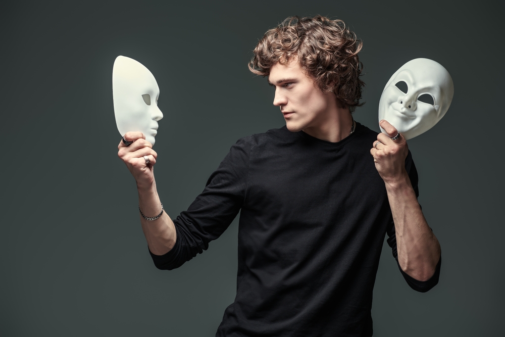 Un jeune homme aux cheveux bouclés, vêtu d’un t-shirt noir, tient deux masques blancs, un dans chaque main. Il regarde intensément le masque dans sa main gauche, au visage neutre, tandis que l’autre masque, dans sa main droite, affiche un sourire théâtral. L’image symbolise le dilemme de l’identité, le choix entre différentes facettes de soi ou les masques que l’on porte dans la vie sociale ou professionnelle.