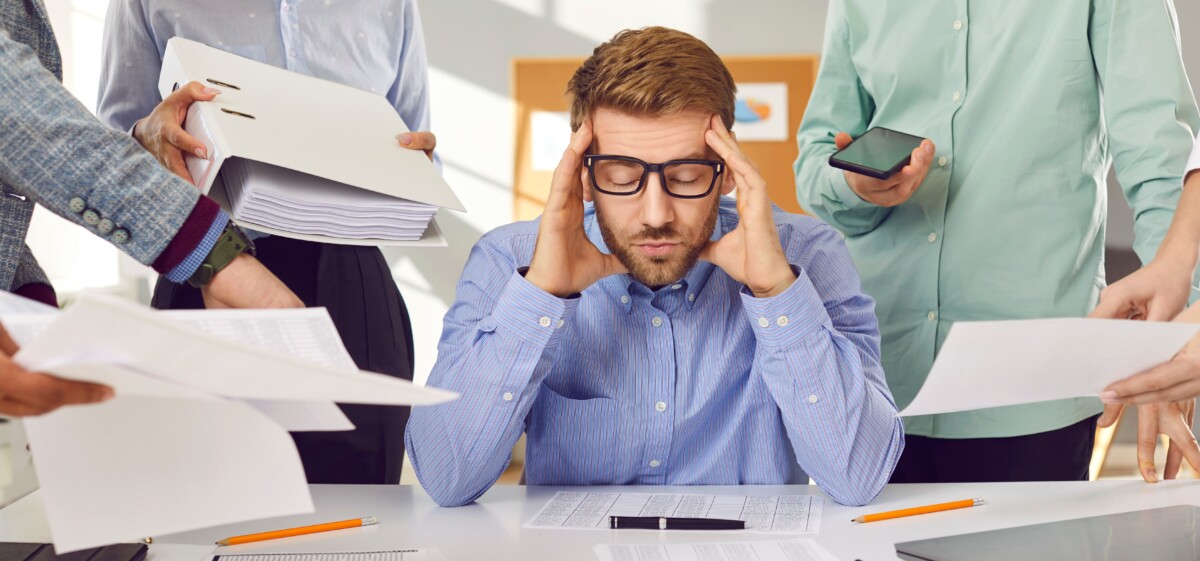 Un manager assis à son bureau, submergé par les demandes, garde les yeux fermés et se prend la tête entre les mains, visiblement dépassé. Autour de lui, plusieurs collègues lui tendent simultanément des dossiers, des documents et un téléphone. L’image illustre une direction ou un management passif, épuisé ou démuni face à des tensions internes, notamment face à un délégué syndical dont les actions ou la posture génèrent des difficultés non traitées. Ce visuel symbolise le manque de préparation ou d’outillage pour faire face à un délégué syndical influent ou stratégique, ce qui alimente frustration et immobilisme.