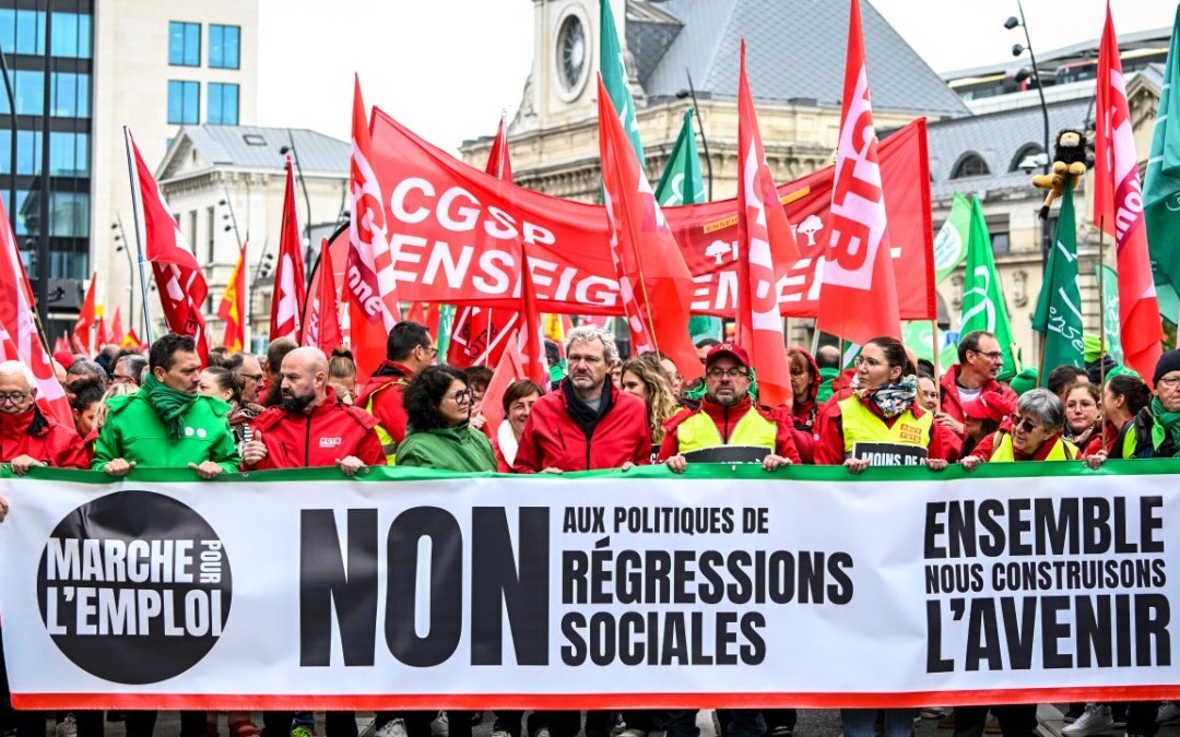 Des manifestants défilent dans une grande ville belge lors d’une grève nationale, portant des drapeaux rouges et verts représentant différentes organisations syndicales. Au premier plan, une large banderole proclame « NON aux politiques de régressions sociales – Ensemble, nous construisons l’avenir ». Cette image symbolise l’unité syndicale et la mobilisation collective autour des enjeux sociaux et économiques. Elle illustre l’article sur la grève du 14 octobre 2025 en Belgique, présentée comme un révélateur du climat social et du dialogue entre employeurs, travailleurs et délégués syndicaux.