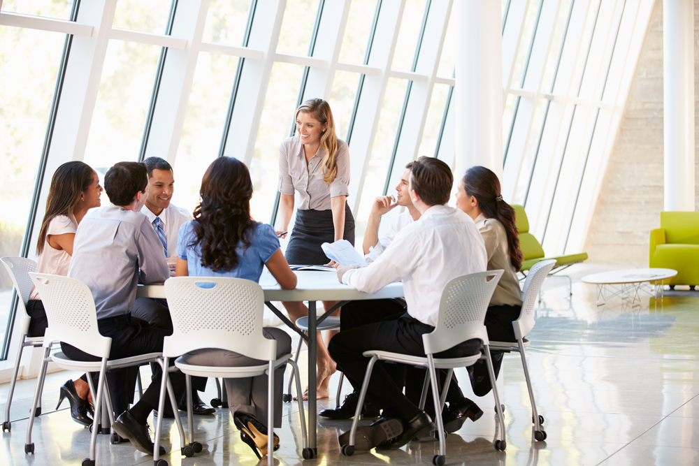 Photographie professionnelle d’une réunion dans une entreprise lumineuse et moderne. Huit collaborateurs sont rassemblés autour d’une table en verre dans une pièce aux grandes baies vitrées. Une femme debout, visiblement responsable ou manager, s’adresse avec bienveillance au groupe assis, qui l’écoute avec attention, certains souriant. L’atmosphère est ouverte, constructive et respectueuse. Cette image illustre l’idée d’un dialogue social sain, fondé sur l’écoute mutuelle, la confiance et la clarté des rôles au sein de l’entreprise.