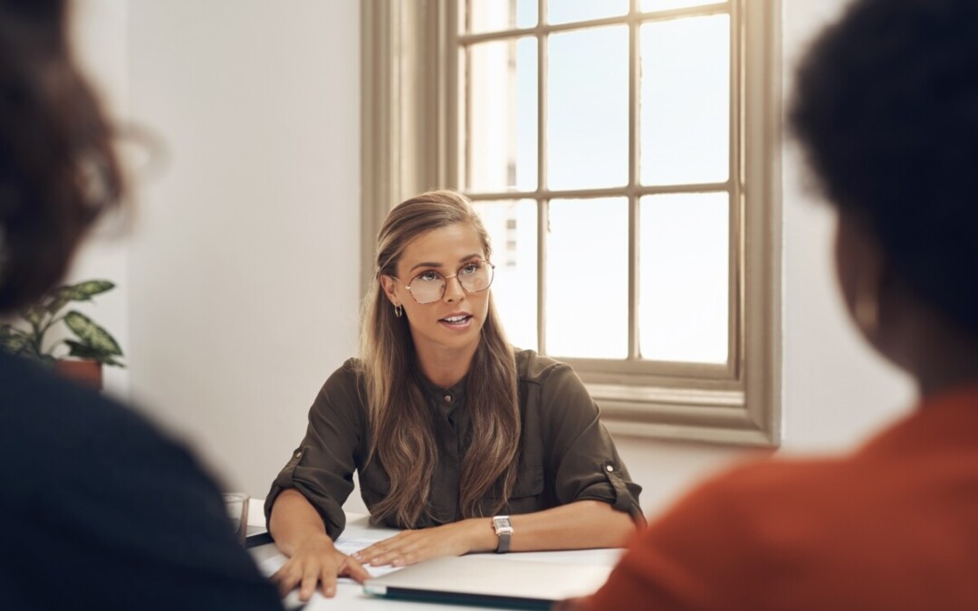 Une consultante ou médiatrice professionnelle, assise face à deux personnes partiellement visibles, anime une réunion dans un bureau lumineux. Elle adopte une posture ouverte, avec une expression attentive et posée, évoquant l'écoute active et l’impartialité. Cette scène illustre une situation d’accompagnement dans un conflit en entreprise, où un expert externe intervient pour faciliter le dialogue, clarifier les enjeux et aider les parties à trouver une issue constructive. L’ambiance suggère à la fois sérieux, professionnalisme et volonté de résolution apaisée.