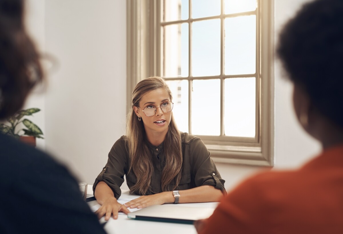 Une consultante ou médiatrice professionnelle, assise face à deux personnes partiellement visibles, anime une réunion dans un bureau lumineux. Elle adopte une posture ouverte, avec une expression attentive et posée, évoquant l'écoute active et l’impartialité. Cette scène illustre une situation d’accompagnement dans un conflit en entreprise, où un expert externe intervient pour faciliter le dialogue, clarifier les enjeux et aider les parties à trouver une issue constructive. L’ambiance suggère à la fois sérieux, professionnalisme et volonté de résolution apaisée.