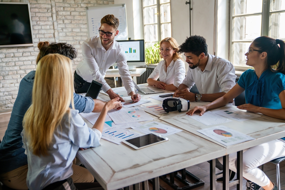 Photo d’un groupe de six jeunes professionnels réunis autour d’une table de réunion, dans une salle lumineuse au mur de briques. L’ambiance est collaborative et positive : une personne debout présente un document aux autres, qui écoutent attentivement en souriant. Des graphiques, rapports et outils numériques sont étalés sur la table (tablette, casque de réalité virtuelle, laptop). L’image illustre un moment de concertation sociale constructive en entreprise, symbolisant l’engagement collectif, le dialogue actif et la recherche de solutions partagées entre acteurs internes.