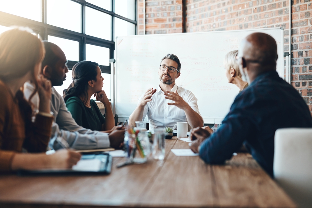 Une réunion professionnelle rassemblant six personnes autour d’une table de travail dans une salle baignée de lumière naturelle. Un homme au centre, visiblement en position de leadership, s’exprime avec conviction pendant que les autres l’écoutent attentivement. En arrière-plan, un tableau blanc présente des schémas stratégiques. L’image symbolise la nécessité de discussions structurées et préparées, notamment lorsqu’une entreprise atteint un seuil critique imposant l’institution d’une délégation syndicale en Belgique. Elle reflète également la posture attendue du management: écoute, anticipation et cadrage.