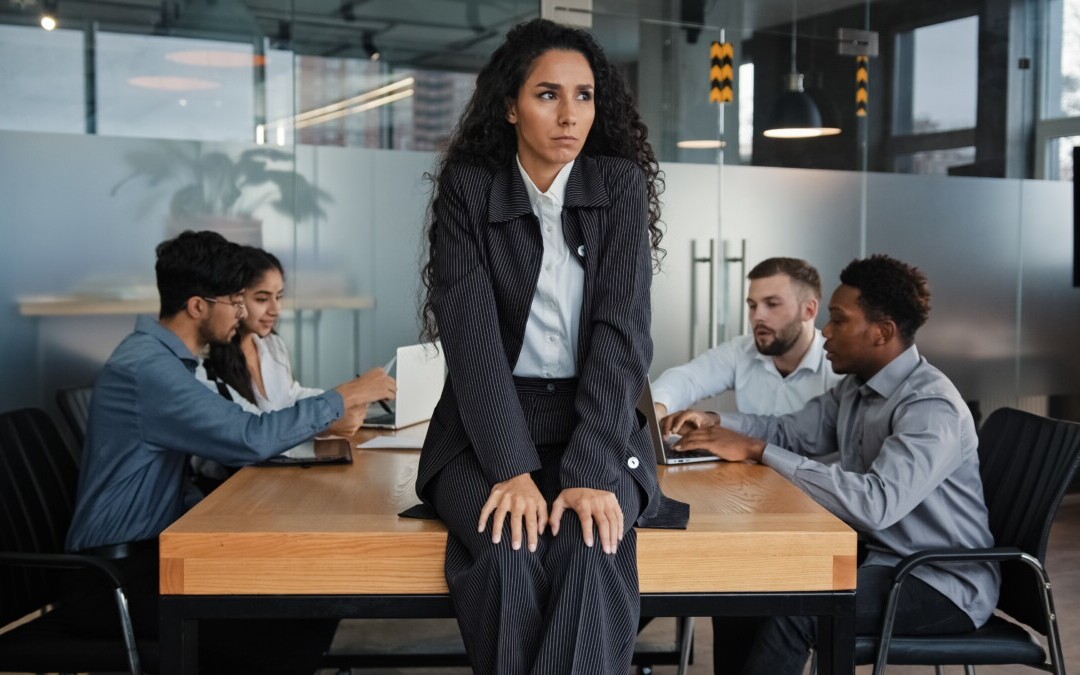 Une femme en costume sombre, au regard sérieux et préoccupé, est assise sur une table de réunion dans un bureau moderne vitré. En arrière-plan, quatre collègues discutent ensemble, l'ignorant visiblement. Cette scène traduit visuellement les tensions sociales, la solitude potentielle du délégué syndical ou la marge d’isolement d’un manager en conflit social. L’image illustre parfaitement le thème de la protection et du licenciement du délégué syndical, en évoquant les impacts humains et relationnels d’un tel processus. Elle souligne l’importance d’un accompagnement stratégique et d’un cadre rigoureux pour gérer ces situations sensibles.
