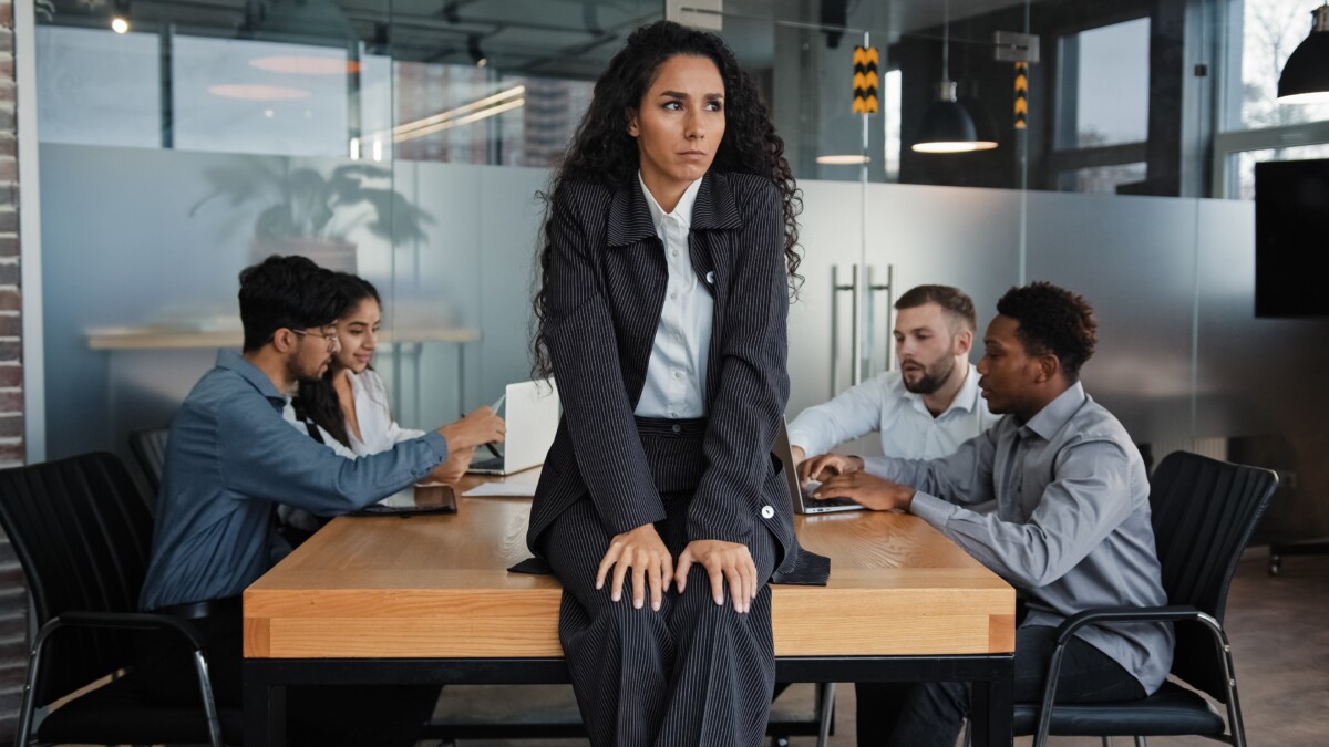 Une femme en costume sombre, au regard sérieux et préoccupé, est assise sur une table de réunion dans un bureau moderne vitré. En arrière-plan, quatre collègues discutent ensemble, l'ignorant visiblement. Cette scène traduit visuellement les tensions sociales, la solitude potentielle du délégué syndical ou la marge d’isolement d’un manager en conflit social. L’image illustre parfaitement le thème de la protection et du licenciement du délégué syndical, en évoquant les impacts humains et relationnels d’un tel processus. Elle souligne l’importance d’un accompagnement stratégique et d’un cadre rigoureux pour gérer ces situations sensibles.
