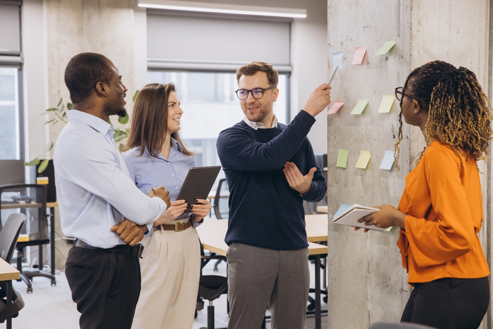 Groupe de professionnels échangeant de manière collaborative autour d’un mur de travail avec post‑it, dans un environnement de bureau moderne. La scène illustre une démarche de Médiation sociale Belgique, visant à prévenir les tensions, structurer le dialogue social et dépasser les blocages avec les délégués syndicaux par des méthodes de concertation et de facilitation adaptées.