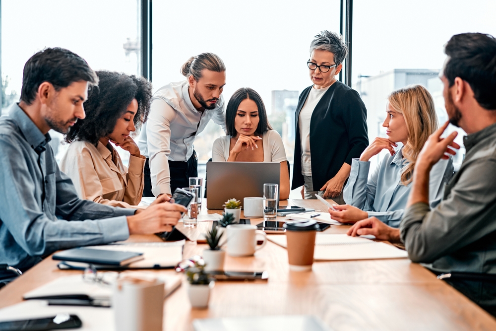 Comprendre le fonctionnement de la délégation syndicale en entreprise en Belgique. Une équipe de collaborateurs diversifiée réunie autour d'une table de conférence en bois dans un bureau lumineux. Au centre, une femme consulte un ordinateur portable pendant que ses collègues, hommes et femmes d'âges différents, observent l'écran avec attention ou discutent. Des tasses de café, des carnets et des tablettes sont disposés sur la table. Cette scène illustre une réunion de concertation au sein d'une délégation syndicale en entreprise en Belgique.