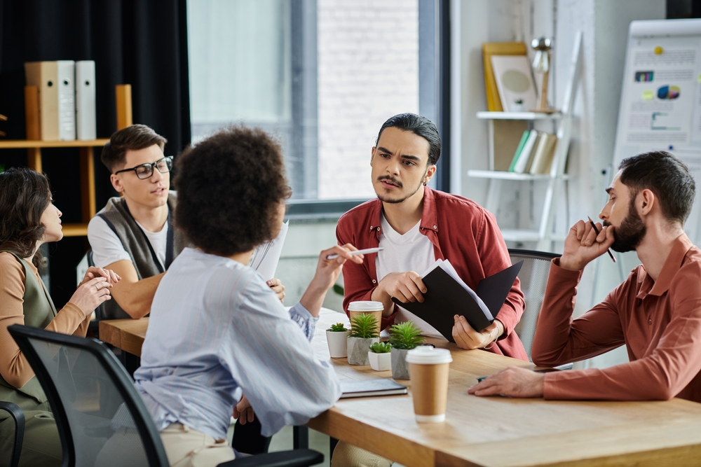 Une réunion d'équipe tendue dans un bureau moderne. Cinq collaborateurs sont assis autour d'une table en bois. Au centre, un homme vêtu d'une chemise rouge, tenant un dossier noir, affiche une expression de confusion et de préoccupation. Une femme de dos, au premier plan, semble argumenter avec insistance en pointant un stylo vers lui. Les autres participants observent la scène avec sérieux et concentration. L'atmosphère suggère une confrontation verbale ou une divergence de vues, illustrant les prémices d'un conflit avec un délégué syndical où la tension émotionnelle commence à prendre le pas sur l'échange technique.
