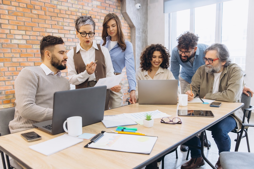 Une équipe multigénérationnelle et diversifiée de six professionnels réunis autour d'une table de réunion en bois dans un bureau de style industriel avec un mur en briques. Au centre, une femme aux cheveux courts et gris pointe un document tout en discutant avec un collègue assis devant un ordinateur portable. À droite, un homme plus âgé avec une barbe grise prend des notes, tandis que d'autres collaborateurs observent un écran de laptop et échangent. Cette scène illustre la transmission des savoirs, la collaboration et la préparation de la relève, des éléments essentiels pour l'avenir du délégué syndical en entreprise.